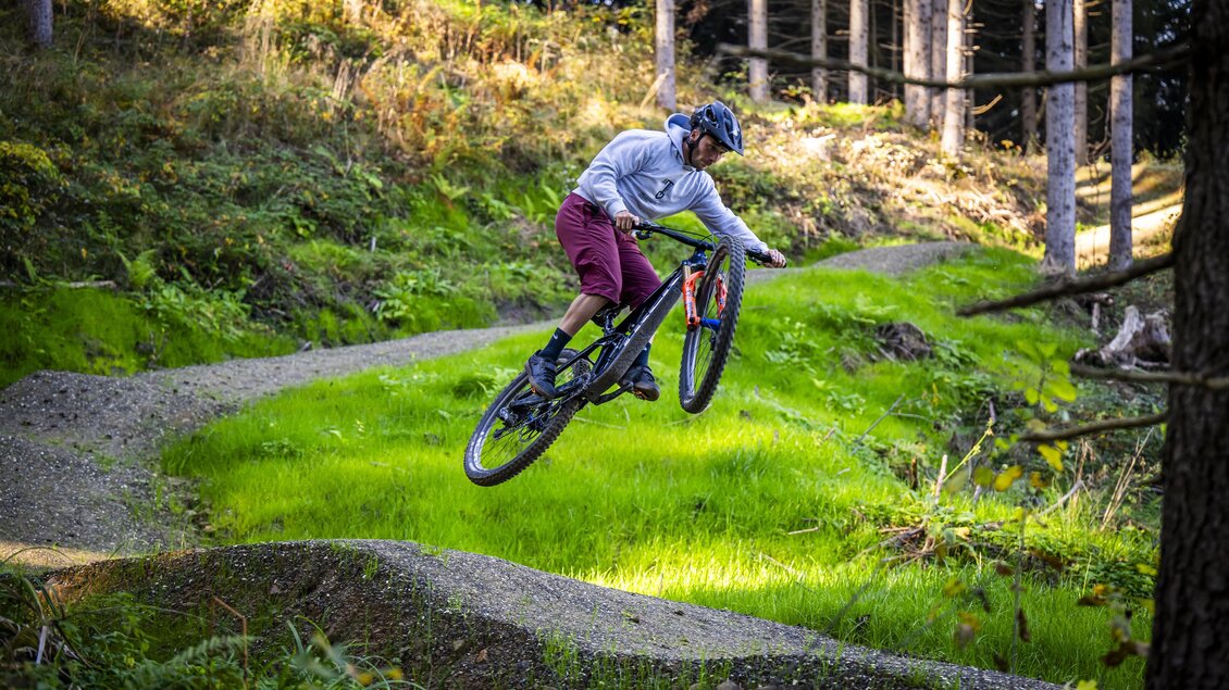 Ein Radfahrer springt über eine Erhebung in einem Waldgebiet. Die Umgebung ist grün und von Bäumen umgeben. | © Lucas Pripfl