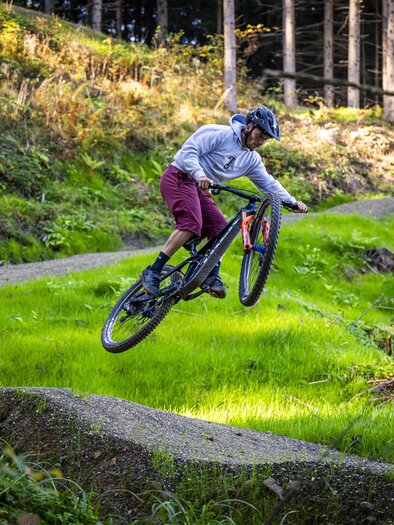 Ein Radfahrer springt über eine Erhebung in einem Waldgebiet. Die Umgebung ist grün und von Bäumen umgeben. | © Lucas Pripfl
