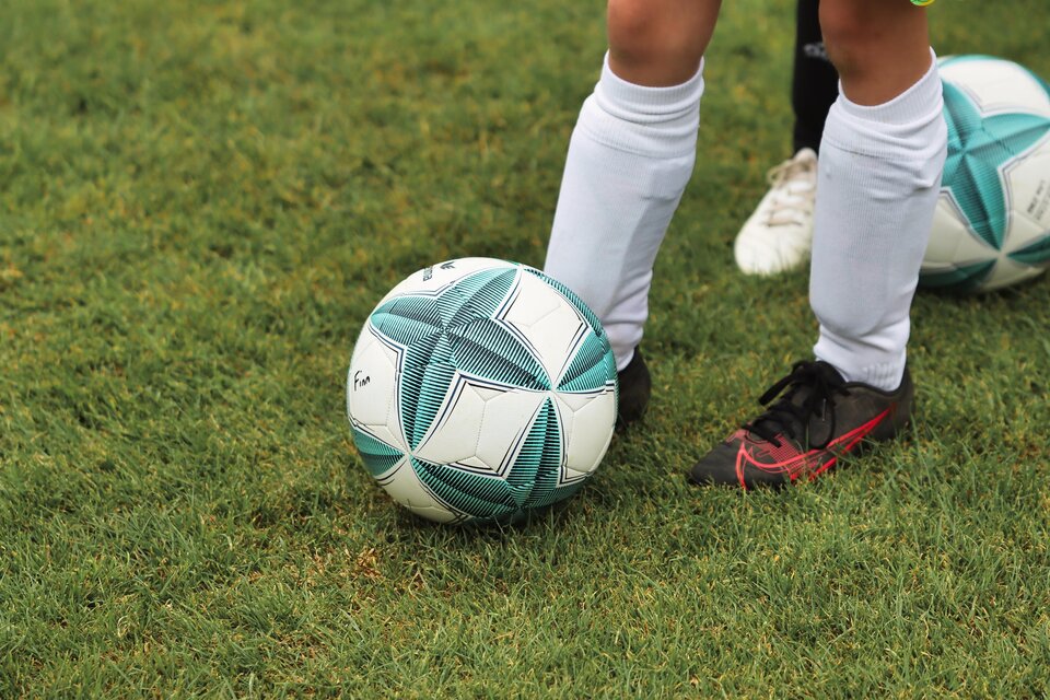 Ein Fußball liegt auf dem Rasen. Im Hintergrund sind die Füße von Spielern in Sportkleidung zu sehen. | © Pixabay