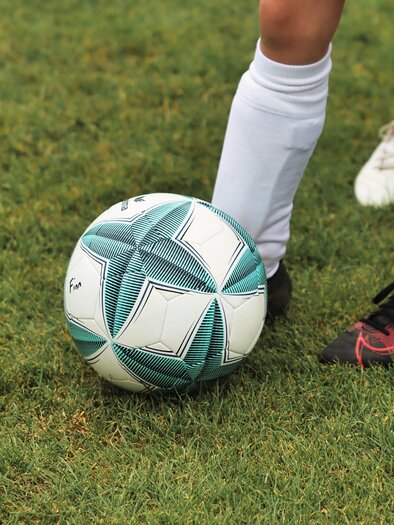 A soccer ball is on the grass. In the background, the feet of players in sportswear can be seen. | © Pixabay