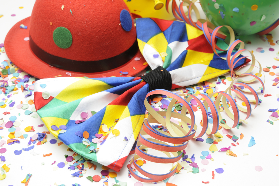 Verschiedene Faschingsartikel wie eine bunt gemusterte Fliege und ein roter Hut, dekoriert mit bunten Luftschlangen und buntem Konfetti auf weißem Untergrund. | © Canva