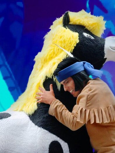 A person is hugging a large, plush horse with a yellow mane. The background is colorful and cheerfully designed. | © Bild: Theater mit Horizont