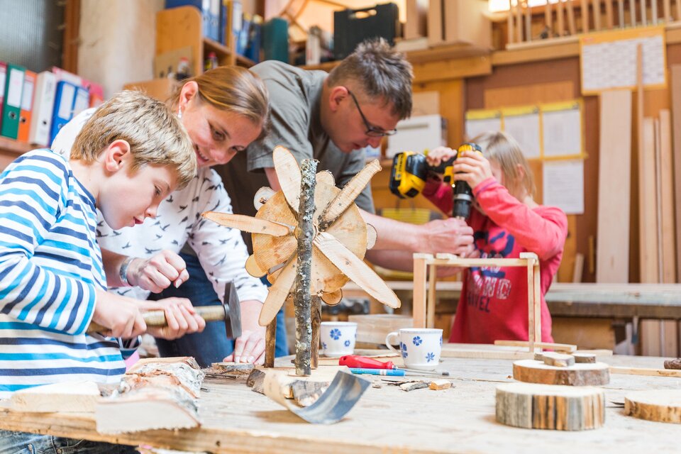 Eine Familie arbeitet gemeinsam an handwerklichen Projekten in einer Werkstatt. Kinder und Erwachsene verwenden verschiedene Werkzeuge, um kreative Holzarbeiten zu erstellen. | © Dominik Steiner