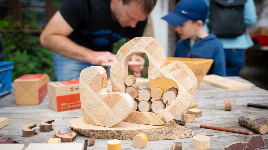 Ein Holztisch mit verschiedenen Holzfiguren, darunter zwei große herzförmige Skulpturen, die mit kleinen Holzstücken gefüllt sind. Im Hintergrund arbeiten ein Erwachsener und ein Kind an Holzprojekten. | © TVB/Laura Passenbrunner
