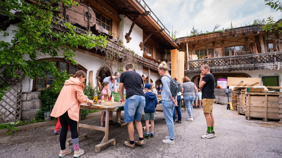 Eine Gruppe von Erwachsenen und Kindern arbeitet an einem Tisch im Freien. Im Hintergrund sind traditionelle Gebäude und Pflanzen zu sehen. | © TVB/Laura Passenbrunner