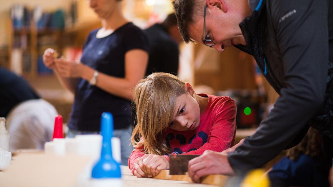 Ein Vater und sein Kind arbeiten gemeinsam an einem Projekt in einer Werkstatt. Im Hintergrund sind weitere Personen mit verschiedenen Materialien beschäftigt. | © Dominik Steiner