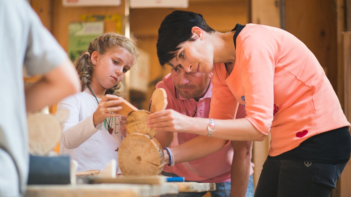 Eine Frau und ein Mädchen arbeiten an einem Holzprojekt. Ein Mann beobachtet sie dabei. | © Dominik Steiner