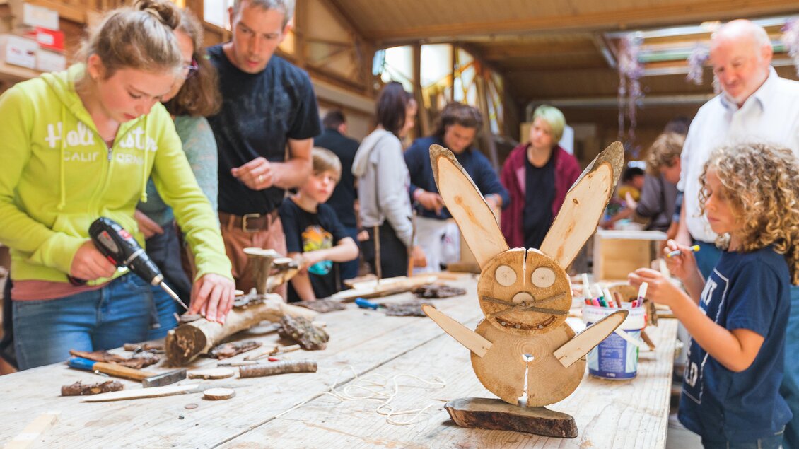 Eine Gruppe von Menschen arbeitet gemeinsam an Holzbasteleien. Im Vordergrund steht ein niedlicher, selbstgemachter Hase aus Holz. | © Dominik Steiner