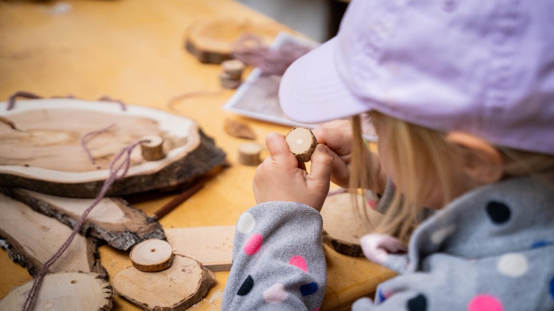 Ein Kind bastelt mit Holzscheiben auf einem Tisch. Es hält ein kleines Stück Holz und schaut konzentriert darauf. | © TVB/Laura Passenbrunner