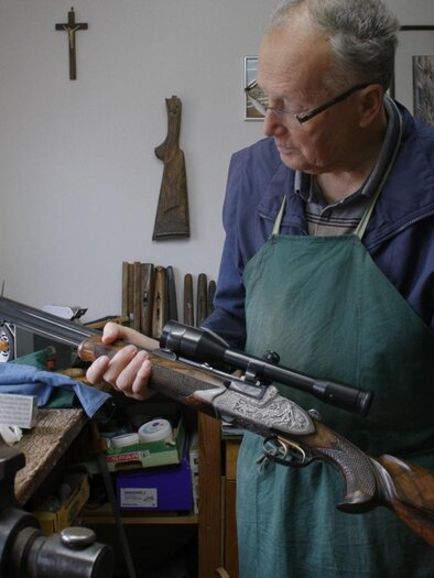Ein älterer Mann in einer Werkstatt hält ein Gewehr in der Hand. Um ihn herum sind verschiedene Werkzeuge und Holzarbeiten zu sehen. | © Sixpackfilm