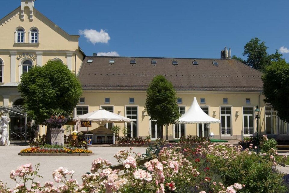 <p>Eine sommerliche Außenansicht des gelben Kur & Congresshaus in Bad Aussee. Vor dem Gebäude befindet sich der Kurpark mit blühenden Rosenbeeten und zwei aufgespannten, weißen Schirmen.  | © TVB Ausseerland - Salzkammergut_Rast