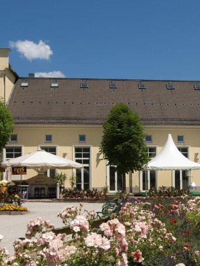 Das schöne Gebäude des Kur- und Congresshaus in Bad Aussee mit gelber Fassade und großen Fenstern in einer sommerlichen Landschaft. Der davor liegende Kurpark ist von einer blühenden Gartenlandschaft umgeben. Unter großen Bäumen stehen Schirme und Tische im Freien. | ©  TVB Ausseerland Salzkammergut_Rastl