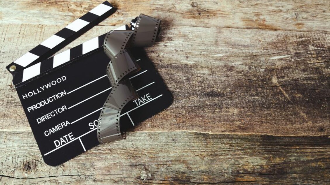 Eine Filmklappe liegt auf einem Holztisch, umgeben von Filmstreifen. Die Klappe zeigt die typischen Hollywood-Symbole. | © freepik