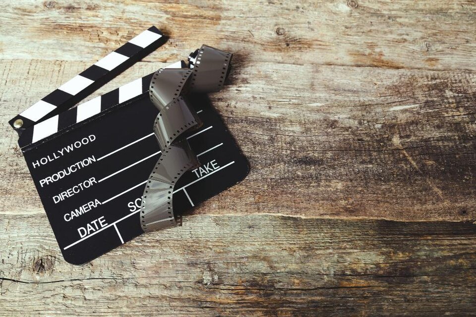 Ein Filmklappe mit einem Filmstreifen liegt auf einer Holztischoberfläche. Die Klappe zeigt typische Beschriftungen für die Filmproduktion. | © Bild: freepik