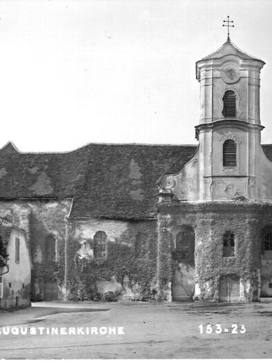 **Historische Aufnahme der Augustinerkirche Fürstenfeld:**
Das Schwarz-Weiß-Foto zeigt die ehemalige Augustinerkirche in Fürstenfeld mit ihrer barocken Fassade und dem markanten Kirchturm. Efeu bedeckt große Teile der Kirchenmauern und verleiht dem historischen Bauwerk einen besonderen Charme. Die ruhige Platzsituation sowie die umliegenden Gebäude vermitteln einen eindrucksvollen Einblick in das Stadtbild vergangener Zeiten. | © Museum Pfeilburg