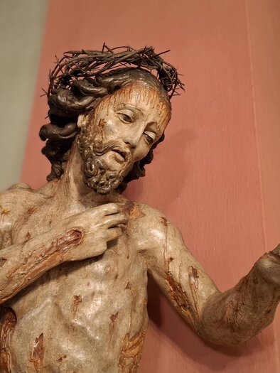 Eine Skulptur von Jesus Christus mit Dornenkrone, die einen nachdenklichen Ausdruck zeigt. Die Figur steht vor einer farbigen Wand in einem Kirchenraum. | © Beate Koller