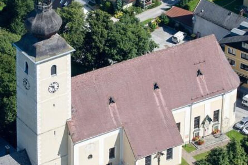 Eine Kirche mit einem hohen Turm und einem roten Satteldach. Umgeben von Wohnhäusern und Bäumen. | © Pfarre Irdning/Harry Schiffer
