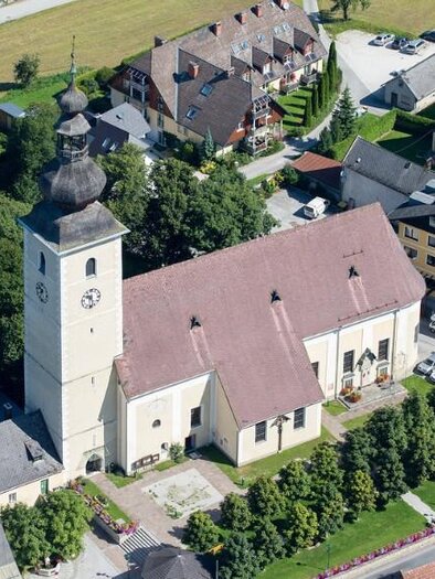 Eine Kirche mit einem hohen Turm und einem roten Satteldach. Umgeben von Wohnhäusern und Bäumen. | © Pfarre Irdning/Harry Schiffer