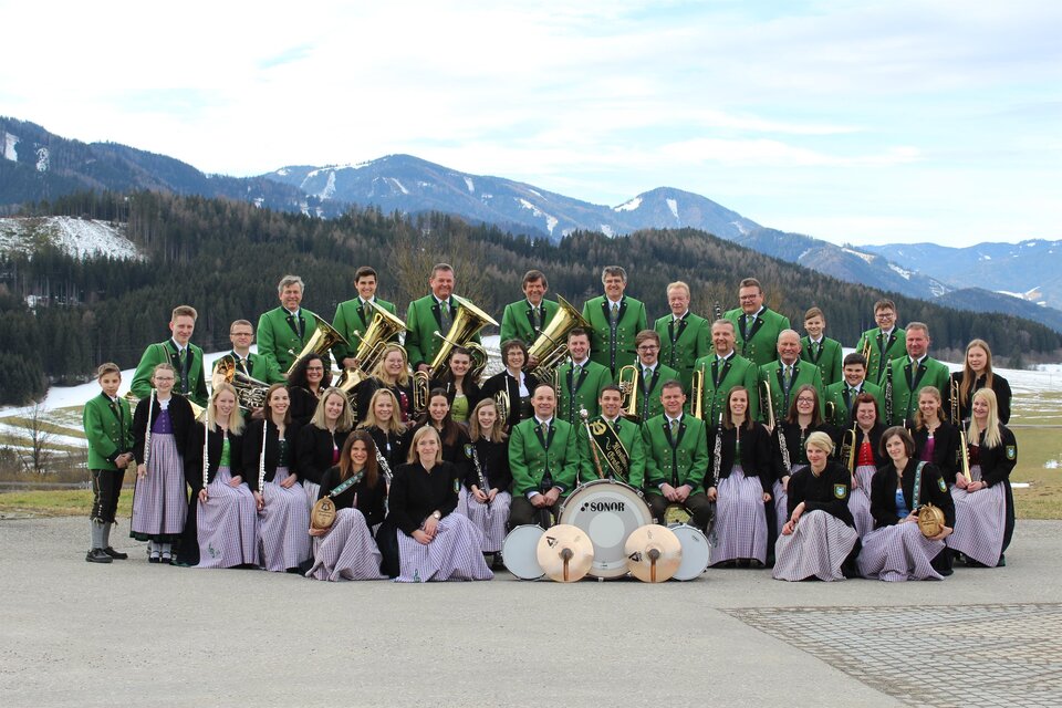 Eine große Musikgruppe in traditionellen Trachten steht vor einer malerischen Berglandschaft. Die Musiker halten Instrumente und lächeln in die Kamera. | © Musikverein Graßnitz