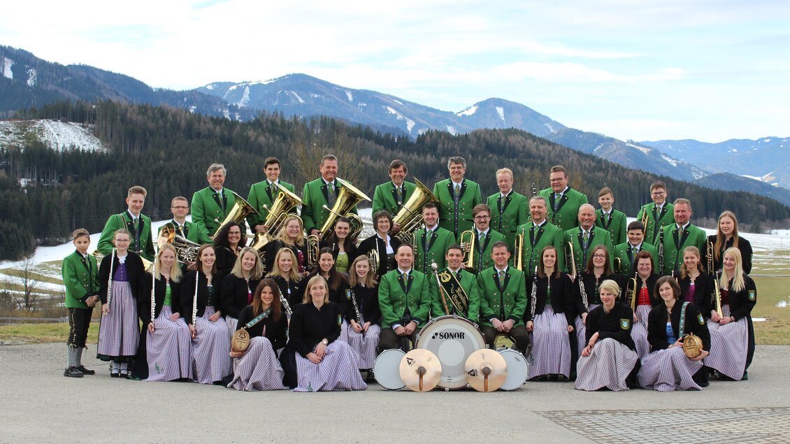 Eine große Musikgruppe in traditionellen Trachten steht vor einer malerischen Berglandschaft. Die Musiker halten Instrumente und lächeln in die Kamera. | © Musikverein Graßnitz