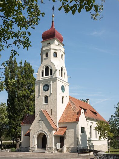 Eine weiße Kirche mit einem roten Turm und buntem Dach in einer grünen Umgebung. Der klare Himmel sorgt für eine ruhige Atmosphäre. | © Foto Schleich