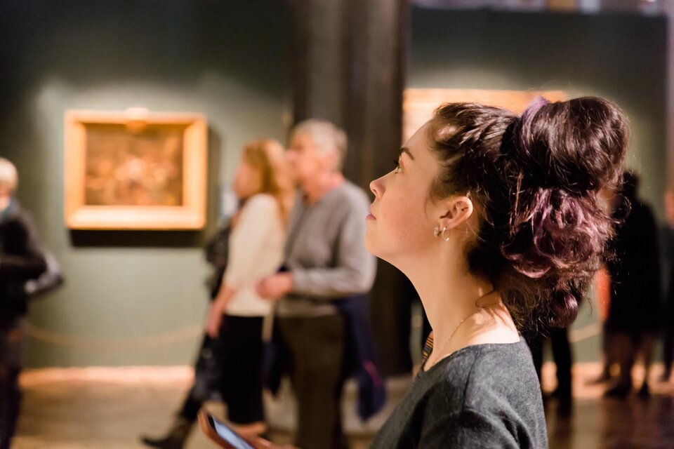 Eine Frau schaut nachdenklich auf ein Kunstwerk in einem Museum. Im Hintergrund sind weitere Besucher zu sehen, die die Ausstellung erkunden. | © AdobeStock