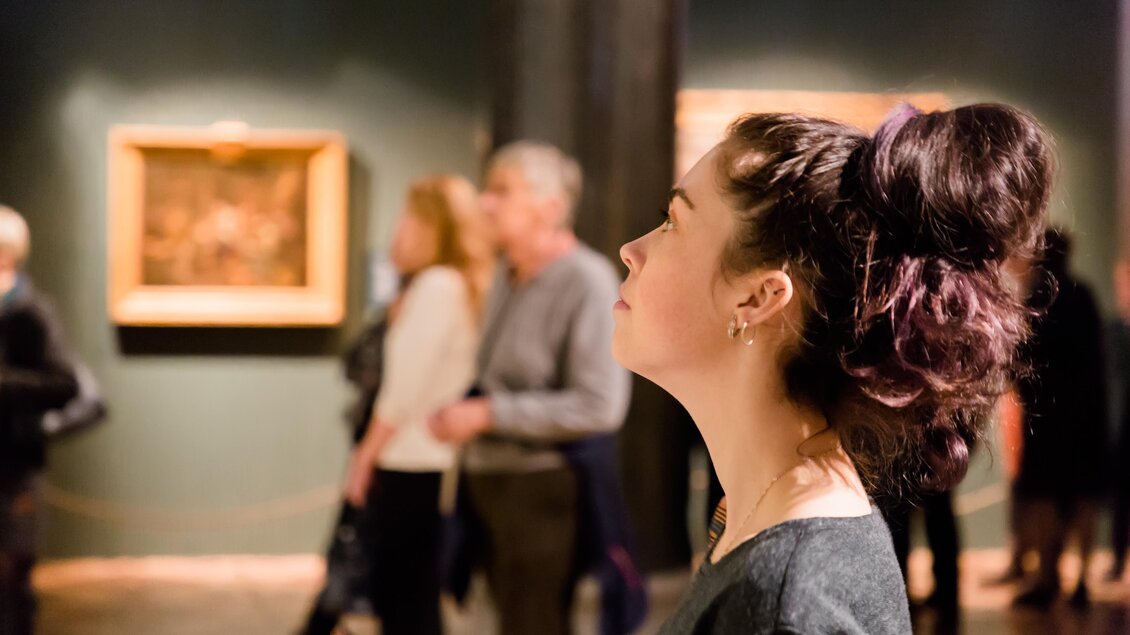 Eine Frau schaut nachdenklich auf ein Kunstwerk in einem Museum. Im Hintergrund sind weitere Besucher zu sehen, die die Ausstellung erkunden. | © AdobeStock