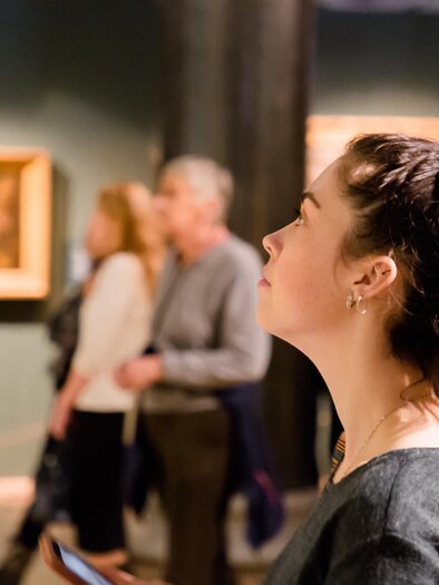 Eine Frau schaut nachdenklich auf ein Kunstwerk in einem Museum. Im Hintergrund sind weitere Besucher zu sehen, die die Ausstellung erkunden. | © AdobeStock