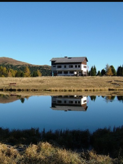 A tranquil lake reflects a large building, surrounded by trees and mountains. The sky is clear and the landscape appears peaceful. | © Andreas Wieser