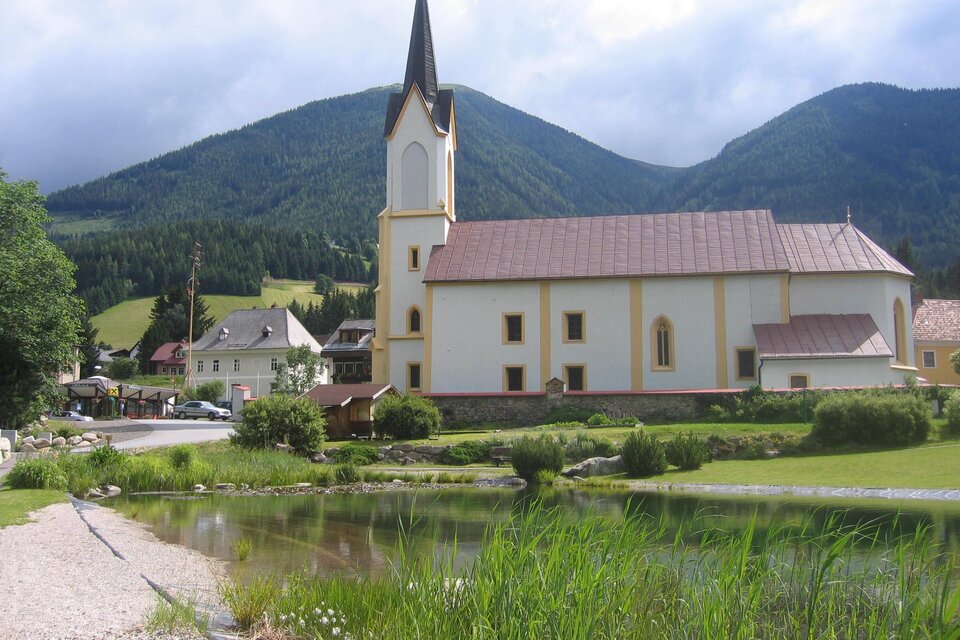 Eine malerische Kirche mit steilem Turm steht am Ufer eines Gewässers. Im Hintergrund sind grüne Hügel und Berge zu sehen. | © Erlebnisregion Murtal