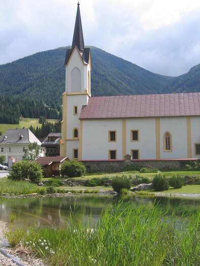 A picturesque church with a steep tower stands on the banks of a body of water. In the background, green hills and mountains can be seen. | © Erlebnisregion Murtal