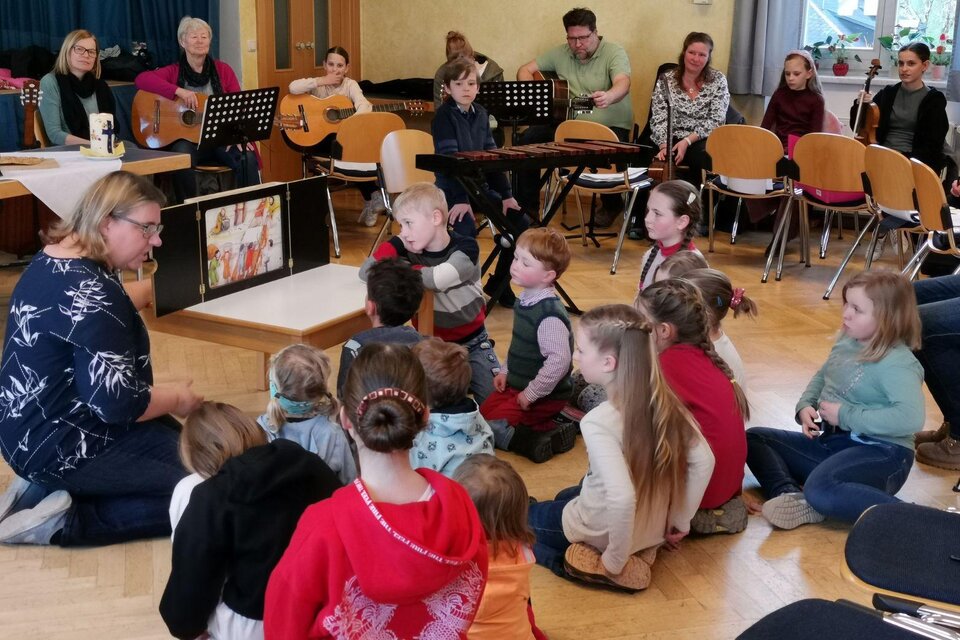 Eine Gruppe von Kindern sitzt im Kreis und hört aufmerksam zu, während eine Frau ihnen etwas erklärt. Im Hintergrund spielen Erwachsene Musik auf Gitarre und sind ebenfalls anwesend. | © (c) Peter Rack, Bad Aussee