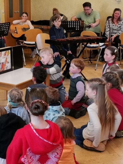 Eine Gruppe von Kindern sitzt im Kreis und hört aufmerksam zu, während eine Frau ihnen etwas erklärt. Im Hintergrund spielen Erwachsene Musik auf Gitarre und sind ebenfalls anwesend. | © (c) Peter Rack, Bad Aussee
