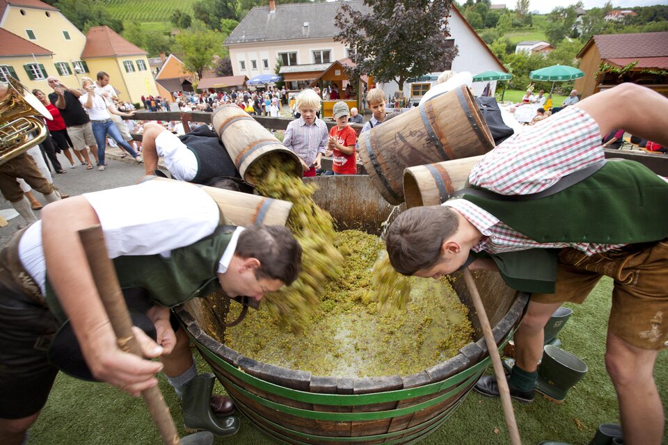 Zwei Männer arbeiten gemeinschaftlich an einem großen Holzfass, in dem eine grüne Masse gerührt wird. Im Hintergrund sind Menschen bei einer Festlichkeit zu sehen. | © TV Thermen- & Vulkanland