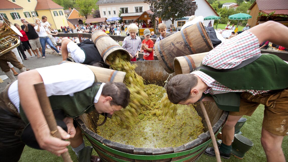 Zwei Männer arbeiten gemeinschaftlich an einem großen Holzfass, in dem eine grüne Masse gerührt wird. Im Hintergrund sind Menschen bei einer Festlichkeit zu sehen. | © TV Thermen- & Vulkanland