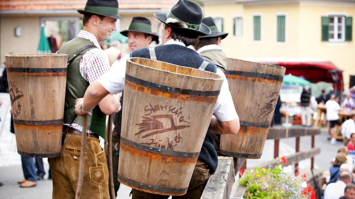 Eine Gruppe von Männern in traditioneller Kleidung trägt Holzbullen. Im Hintergrund sind festliche Stände und eine Menschenmenge zu sehen. | © TV Thermen- & Vulkanland