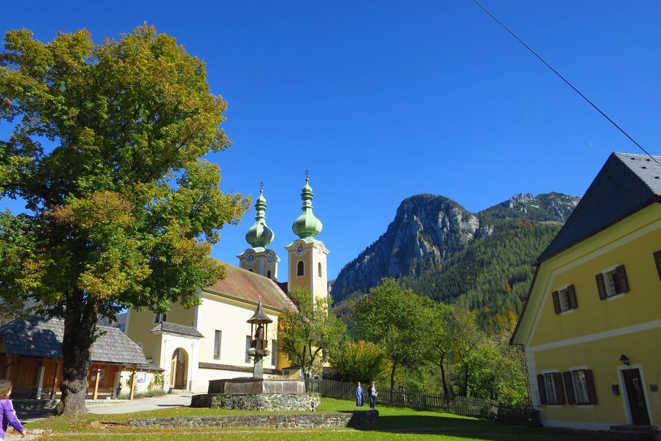 Eine malerische Landschaft mit einem historischen Gebäude und zwei Kirchtürmen. Im Hintergrund erheben sich grüne Berge unter einem klaren blauen Himmel. | © Ludwig Gottsbacher
