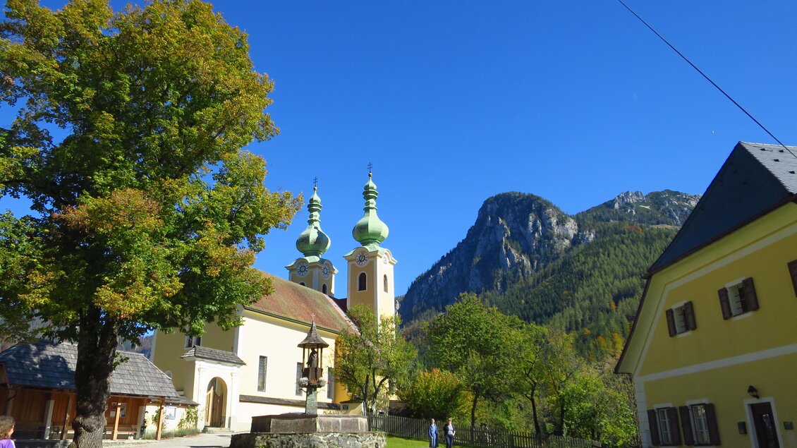 Eine malerische Landschaft mit einem historischen Gebäude und zwei Kirchtürmen. Im Hintergrund erheben sich grüne Berge unter einem klaren blauen Himmel. | © Ludwig Gottsbacher