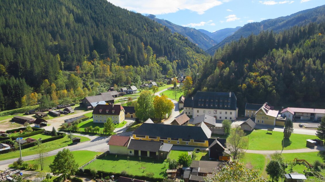 Eine malerische Landschaft mit einem kleinen Dorf in einem Tal. Umgeben von grünen Bergen und klarer blauer Himmel. | © Ludwig Gottsbacher