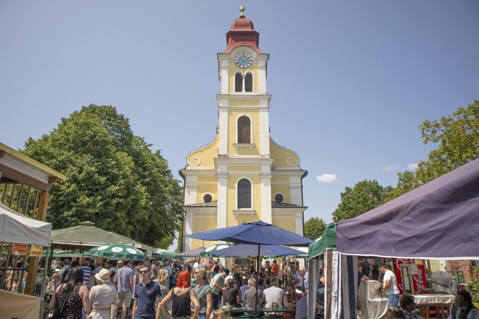 Ein belebter Marktplatz mit vielen Menschen und bunten Marktständen. Im Hintergrund steht eine gelbe Kirche mit einem roten Turm und einer Uhr. | © (c) Thomas Brandl