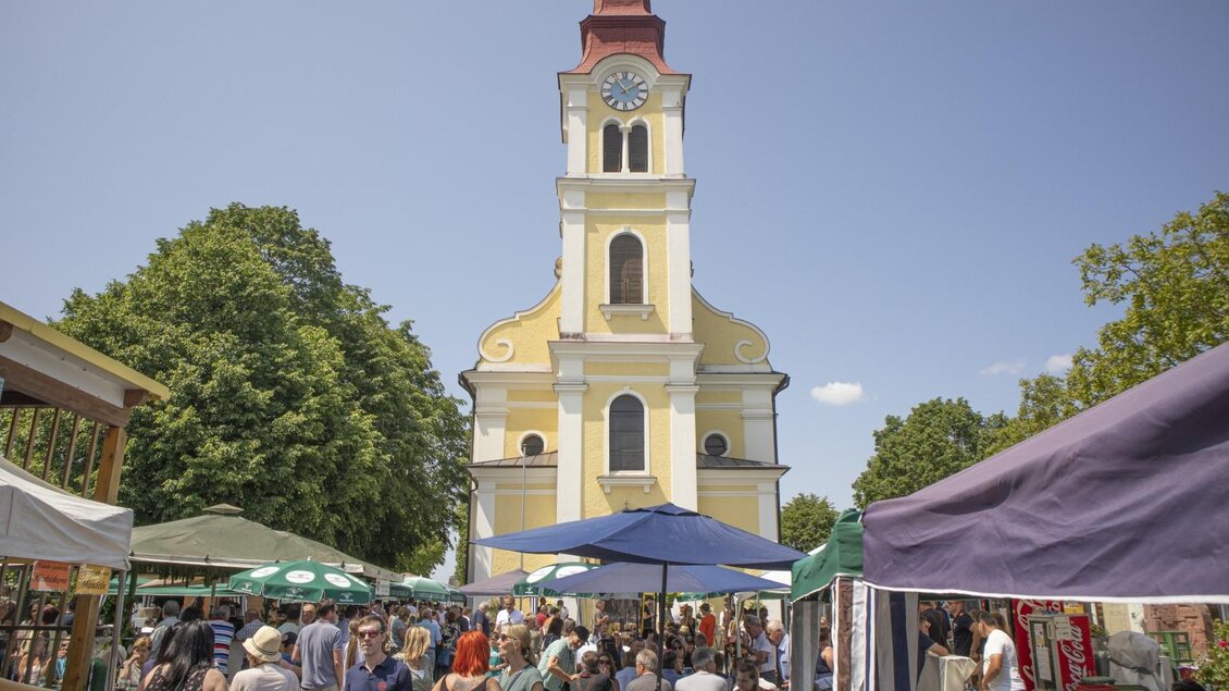 Ein belebter Marktplatz mit vielen Menschen und bunten Marktständen. Im Hintergrund steht eine gelbe Kirche mit einem roten Turm und einer Uhr. | © (c) Thomas Brandl