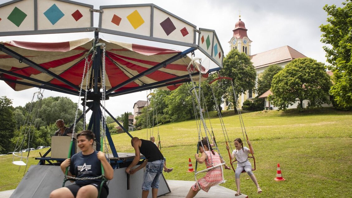 Ein Spielplatz mit einem Karussell und schwingenden Sitzen. Im Hintergrund sieht man eine Kirche und grüne Bäume. | © (c) Thomas Brandl