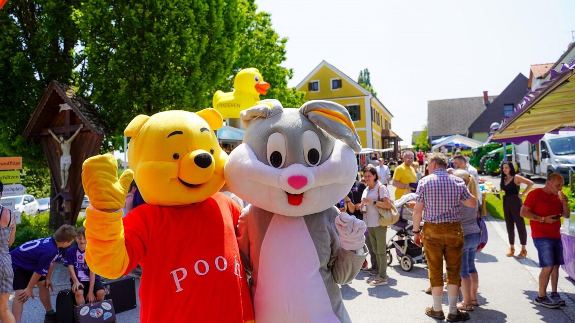 Zwei Maskottchen, ein Bär und ein grauer Hase, posieren lächelnd auf einer belebten Straße. Im Hintergrund sind Menschen und bunte Stände zu sehen. | © (c) Ingrid Donnerer