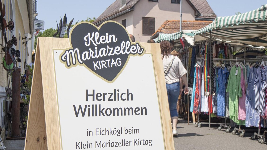 Ein Willkommensschild für den Klein Mariazeller Kirtag. Im Hintergrund sind Verkaufsstände und Menschen zu sehen. | © (c) Thomas Brandl
