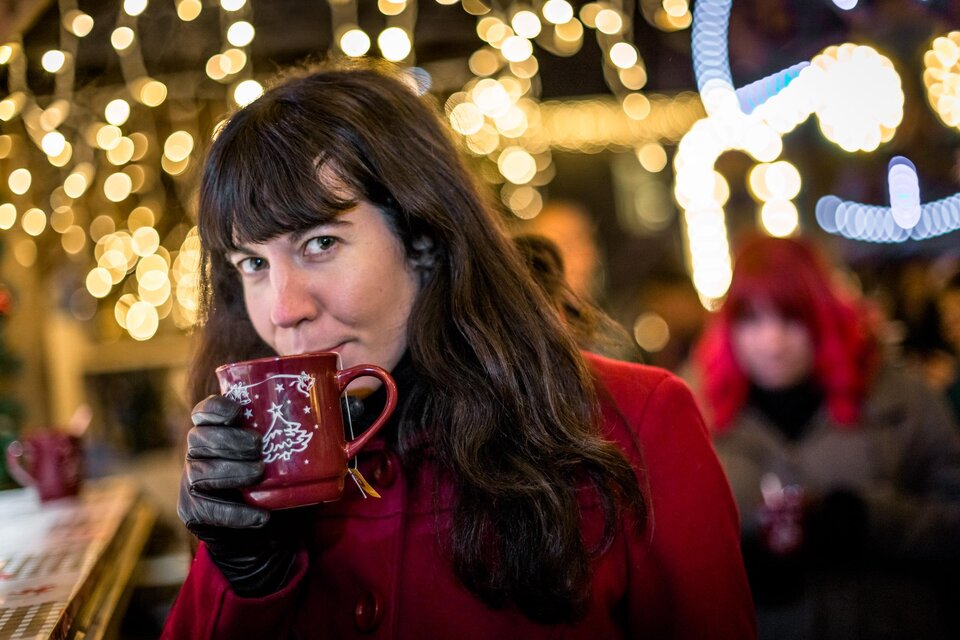Eine Frau mit langen, dunklen Haaren trinkt aus einer roten Tasse und trägt einen roten Mantel. Im Hintergrund sind funkelnde Lichter und weitere Personen zu sehen. | © TV Südsteiermark | Stephan Friesinger