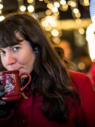 A woman with long, dark hair is drinking from a red cup and is wearing a red coat. In the background, there are sparkling lights and other people visible. | © TV Südsteiermark | Stephan Friesinger