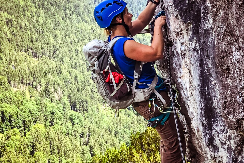 Ein Kletterer in einem blauen Helm klettert an einer Felswand. Im Hintergrund sind grüne Wälder und Berge zu sehen. | © Tourismusverband Schladming-Dachstein