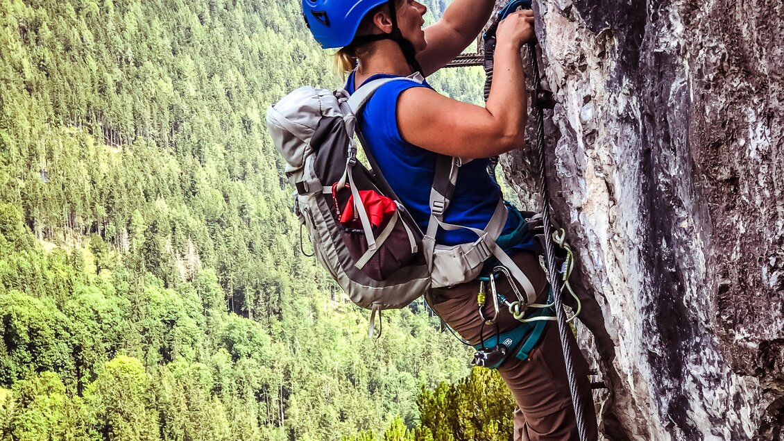 Ein Kletterer in einem blauen Helm klettert an einer Felswand. Im Hintergrund sind grüne Wälder und Berge zu sehen. | © Tourismusverband Schladming-Dachstein