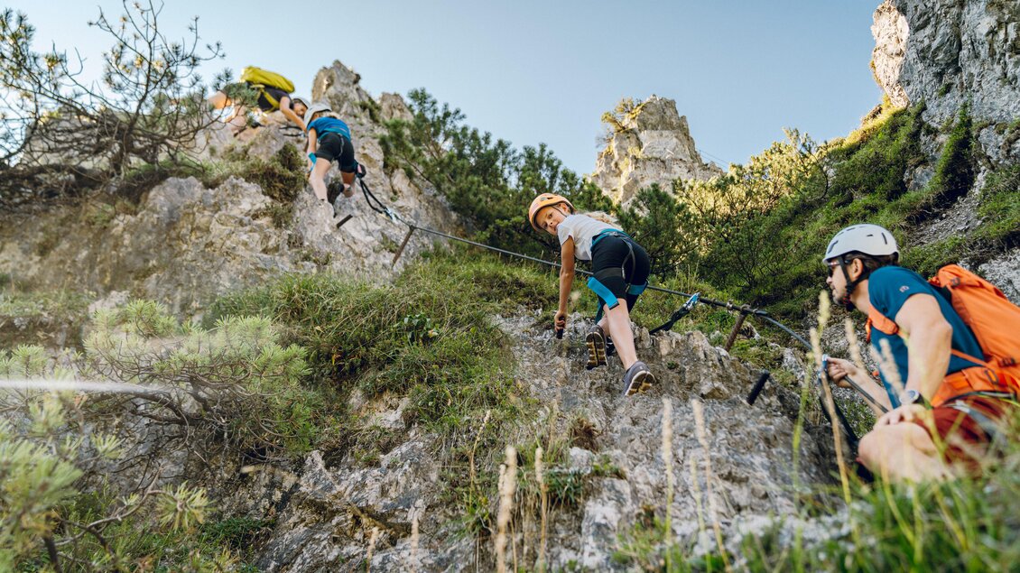 Eine Gruppe von Kletterern erklimmt eine felsige Steigung mit Seilen. Die Landschaft ist grün und sonnig, ideal für Outdoor-Aktivitäten. | © Christine Höflehner