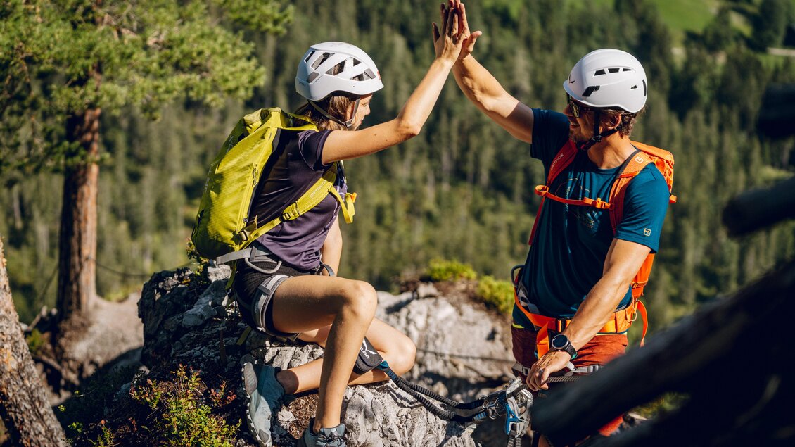 Zwei Bergsteiger geben sich ein High-Five auf einem Felsen. Im Hintergrund sind grüne Wälder und eine bergige Landschaft zu sehen. | © Christine Höflehner